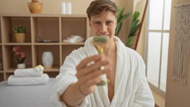 Kaplıca odasında elinde yeşim taşıyla beyaz bornoz giyen genç bir adam. Etrafı huzur ve güzellik ürünleriyle çevrili..