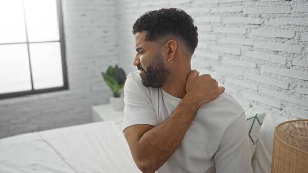 Молодой человек смотрит с дискомфортом, держа плечо в современной спальне с белыми кирпичными стенами и окном, предполагая, что он может испытывать боль или дискомфорт.