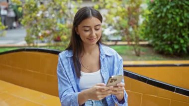 Şehir arka planında akıllı telefon kullanan genç bir kadın çizgili bir gömlek giyip gülümsüyor.