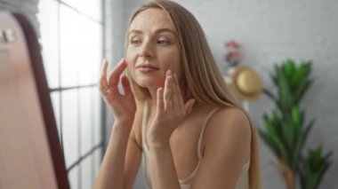 Parlak oturma odasında cilt bakımı yapan bir kadın. Kapalı alandaki bitkilerin yanındaki geniş bir pencereden süzülen doğal ışık. Evde sakin bir atmosfer yayıyor..