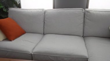 Genç adam, oturma odasında gri bir koltukta turuncu yastıkla oturmuş dizüstü bilgisayarla çalışırken...