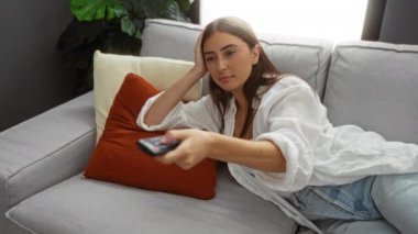 Oturma odasındaki kanepede uzanan bir kadın, uzaktan kumandayı elinde tutuyor, gelişigüzel giyinmiş, arka planda bir bitki ve pencereden içeri giren doğal ışıkla..