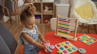 Dövmeli bir anne, kızına oyuncaklarla oyun odasında yardım ediyor, sıcak ve kapalı bir ortamda öğrenme ve bakıma odaklanıyor, aileyi ve yaratıcılığı ön plana çıkarıyor..