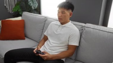 Genç adam oturma odasında bir kanepede oturuyor, kumandayla video oyunları oynuyor, modern, kapalı bir evde gri bir kanepe ve turuncu bir yastık var..