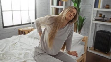 Yatak odasında sırt ağrısı çeken, pijama giyen, omuzlarından dökülen sarı saçlı, modern iç mekan penceresinden görülebilen, iç mekan rahatsızlığı olan bir kadın..