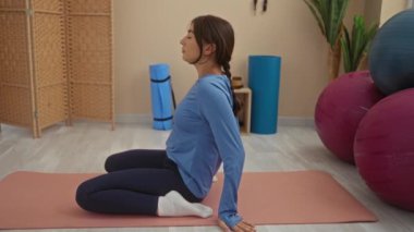 Spor salonunda egzersiz yapan, spor aletleri ve denge toplarıyla çevrili minderde yoga yapan genç bir kadın.