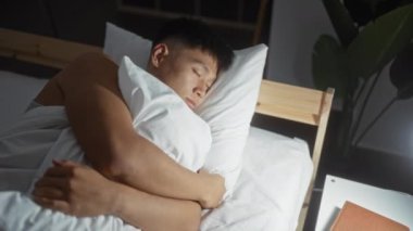 Asyalı bir adam yatak odasında huzurlu bir şekilde uyurken bir yastığa sarılıp, rahat bir ortam hazırlıyor..