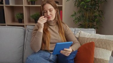 Kadın rahat bir oturma odasında, çiçekli, gözlüklü ve süveterli, rahat ev atmosferi, doğal ışıklandırma, kot pantolon, rahat bir ortam ile rahat bir şekilde otururken tablet okuyor..