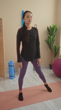 Çinli bir kadın spor salonunda esneme egzersizleri yapıyor. Paspaslar ve egzersiz topları gibi spor malzemeleriyle çevrili. Yoga egzersizleri yapıyor..
