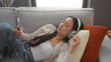 Oturma odasındaki koltukta akıllı telefon ve kulaklıkla dinlenen bir kadın.