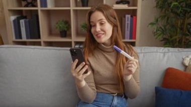 Kadın, rahat oturma odasında videolu yayında hamilelik testini heyecanla tutuyor ve neşeyi olumlu bir şekilde ifade ediyor..