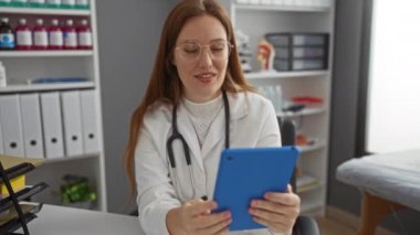 Klinikteki kadın doktor iletişim için tablet kullanıyor, gülümsüyor ve profesyonel bir muayenehanede el sallıyor..