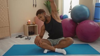 Kapalı bir spor salonundaki mavi yoga minderinin üzerinde uzanan genç adam spor topları, mumlar ve spor aletleriyle sakin bir ortamda.