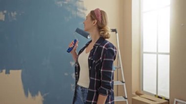 Parlak oturma odasında tekerlekli sandalyeyle duvar boyayan, işine gülümseyen, günlük kıyafetler giyen, kutular ve merdivenlerle çevrili bir kadın, yeni bir başlangıç öneriyor..