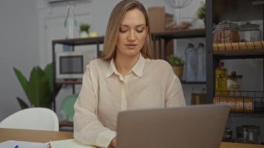 Modern ofis ortamında, sarı saçlı bir kadın dizüstü bilgisayar ve defter kullanarak etrafını bitki ve raflarla çevrili bir masada otururken odak ve rahatlık iletiyor..