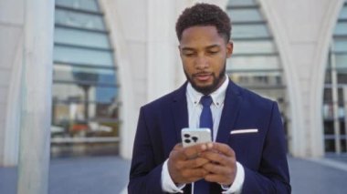 Mavi takım elbiseli genç adam şehir merkezinde akıllı telefon kullanıyor ve arkasında modern mimari var.