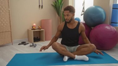 Spor salonundaki yoga minderinde egzersiz yapan genç bir adam. Etrafı spor topları ve minimalist dekorlarla çevrili. Modern, dingin bir ortamda fitness ve sağlığa odaklanıyor..