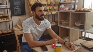 Sakallı genç bir adam arka planda ekmek ile fırında oturuyor. Kol saatine bakıyor. Parlak sarı bir fincandan kahve içiyor.