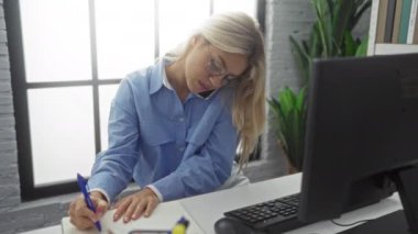 Modern ofiste telefonla konuşurken notlar yazan bir kadın masa başında bir bilgisayar ve arka planda büyük bir pencere, günlük mavi gömlek ve gözlük takıyor..