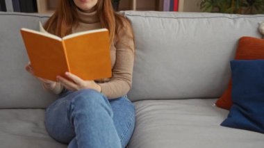 Rahat bir oturma odasında gri bir kanepede dinlenen, rahat bir kot pantolon ve bej bir kazak giyen, renkli yastıklarla çevrili bir kadın..