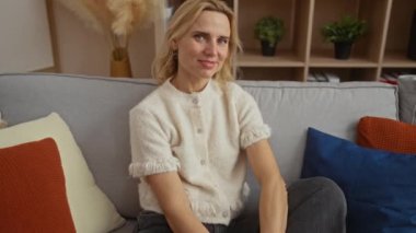 Rahat bir oturma odasında rahat bir kanepede oturan kadın, rahat bir gülümseme sergiliyor, renkli yastıklar ve yeşilliklerle çevrili..