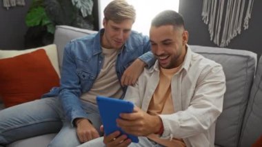 Rahat bir oturma odasında rahatlayan iki adam, tablete bakarken gülümseyen, günlük kıyafetler ve modern ev dekoruyla birlikte kaliteli zaman geçiren iki adam.