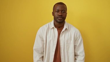 Afro-Amerikan gömlekli adam kollarını kavuşturmuş, kameraya bakıyor ve izole edilmiş sarı arka planda gülümsüyor, elleri çenesinde olumlu düşünüyor.