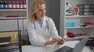 Klinikte steteskoplu beyaz önlüklü bir kadın, iş yerinde tıbbi ekipman ve belgelerle çevrili akıllı telefon kullanıyor.
