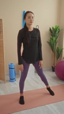 Çinli bir kadın spor salonunda esneme egzersizleri yapıyor. Paspaslar ve egzersiz topları gibi spor malzemeleriyle çevrili. Yoga egzersizleri yapıyor..