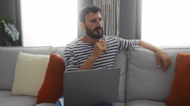 Oturma odasında koltukta oturan sakallı genç adam, evde dizüstü bilgisayarla çalışırken düşünüyor..