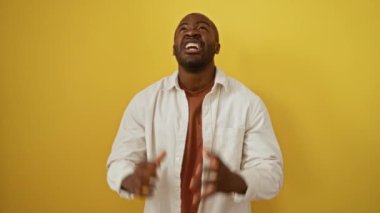 Afrika kökenli Amerikalı bir adam, çılgın bir ifadeyle ayakta duruyor, kollarını kaldırarak bağırıyor, çılgın ve sinirli bir şekilde, izole edilmiş sarı arka planda agresif bir şekilde.