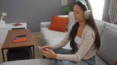 Modern bir oturma odasında, ahşap bir masa ve turuncu yastıkla rahat bir koltukta otururken, kulaklıklı müzikten hoşlanan bir kadın.