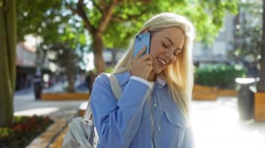 Şehir parkında yeşil ağaçlar ve bulanık şehir arka planında telefonla konuşurken gülümseyen bir kadın..