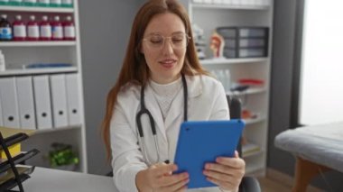 Klinikteki kadın doktor iletişim için tablet kullanıyor, gülümsüyor ve profesyonel bir muayenehanede el sallıyor..
