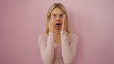Ağzını kapatan şaşkın bir kadın, pembe bir arka plan karşısında duruyor, uzun kollu bir gömlek giyerken sarı saçları ve genç görünümünü sergiliyor.