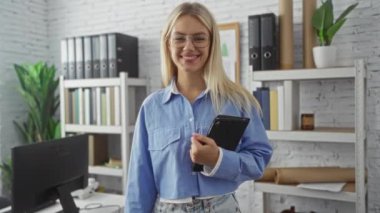 Modern bir ofis ortamında gülümseyen, etrafı kitaplarla ve dekorlarla çevrili bir kadın, diğer elinde tablet tutarken dostça bir el sıkışma teklif ediyor ve gözlüklü günlük kıyafetler giyiyor..