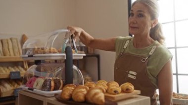 Arka planda çeşitli ekmekler sergilenen kapalı bir pastaneden hamur işi seçen yaşlı kadın.