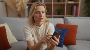 Modern oturma odasındaki rahat koltukta akıllı telefon kullanan bir kadın, sarışın, yetişkin, beyaz bir kadının ev içinde şık bir şekilde etkileşime geçtiği bir ev..