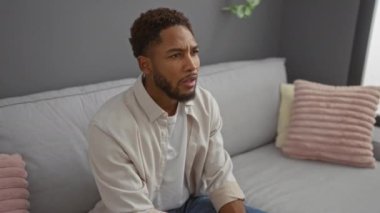 Oturma odasında otururken elini ağzını kapatan genç adam şaşırmış görünüyor.