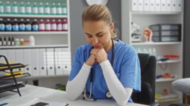 Mavi önlüklü kadın bir klinikte steteskopla oturmuş rafların ve ofis malzemelerinin arasından akıllı telefona bakıyor düşünceli ve odaklanmış görünüyor..