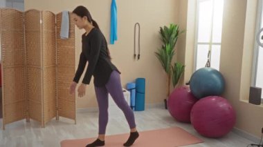 Spor salonundaki paspasın üzerinde yoga yapan bir kadın yoga pozunda denge ve esnekliğe odaklanıyor. Etrafı egzersiz ekipmanları ve bitkilerle çevrili.