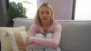 Oturma odasındaki kanepede oturan kadın düşünceli görünüyor, minderler ve bitkilerle çevrili, rahat bir evde rahat kıyafetler giyiyor..