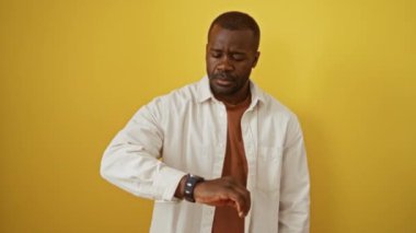 Afro-Amerikalı bir adam gömleğiyle ayakta duruyor. Bilek saatini kontrol ediyor. İzole edilmiş sarı arka planda.