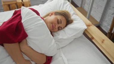 Beyaz yatakta huzur içinde uyuyan bir kadın rahat bir yatak odasında, tahta bir yatak başlığıyla yastığa sarılıyor. Evinde rahatlık ve rahatlık öneriyor.
