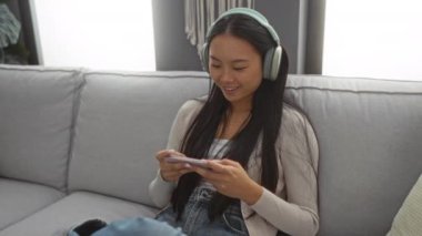 Uzun siyah saçlı genç bir kadın, sıcak aydınlatılmış oturma odasında rahat bir koltukta otururken kulaklık takıyor ve akıllı telefon kullanıyor.