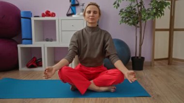 Spor salonunda yoga minderiyle meditasyon yapan bir kadın. Yoga topu ve halterler de dahil olmak üzere, egzersiz sırasında odak dengesini ve rahatlığını vurgulayan halterlerle çevrili..