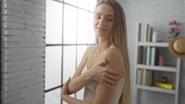 Parlak oturma odasında gülümseyen, sarı saçlı ve geniş pencerenin yanında rahat bir poz veren bir kadın ve güneş ışığının sıcak yansımasıyla iç mekanda rahat bir ortam sunan bir kitaplık..