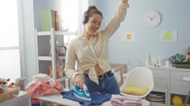 Neşeli bir çamaşırhanede kıyafetlerini ütüleyen, kulaklık takan ve ütü masasının yanında dans eden, katlanmış çamaşır ve oyuncaklarla çevrili bir kadın..