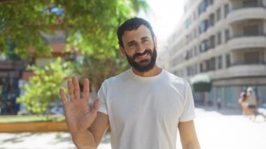 Güneşli bir günde arka planda ağaçlar ve binalar olan şehir parkında el sallayan yakışıklı bir adam.