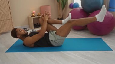 Spor salonunda mavi hasırla egzersiz yapan genç adam arka planda mum ve egzersiz toplarıyla bacak esneme hareketleri yapıyor.